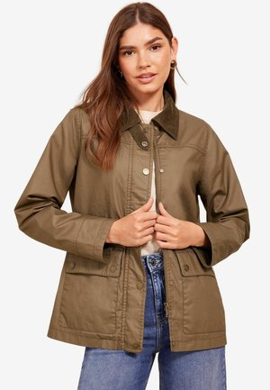 Olivgrüne Jacke mit Cordkragen, Druckknopfverschluss, Brust- und Seitentaschen sowie einer lockeren Passform, getragen über einem beigen Oberteil und blauen Jeans.