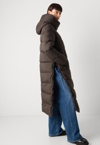 Bruine pufferjas met een lange, doorgestikte ontwerp, capuchon en zijsplitten, gecombineerd met wijde blauwe jeans en zwarte schoenen.