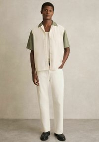 Cămașă cu mânecă scurtă, cu nasturi în crem, cu guler verde, asociată cu pantaloni crem și pantofi negri. Textură netedă, design simplu, detaliu subtil al buzunarului.