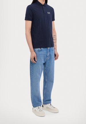 Homme portant un polo bleu marine EA7, un jean bleu clair, des baskets blanches et avant-bras tatoué visible, debout devant un fond uni.