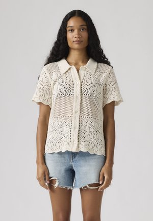 Mujer con cabello largo y rizado, vistiendo una blusa de manga corta color crema con botones y un pantalón corto de mezclilla clara desgastado.