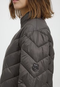 Pilkas pūkinis švarkas su aukštu apykakle, zigzago siuvinėjimu ir logotipu ant peties. Lygus, blizgus paviršius, lengvas dizainas.