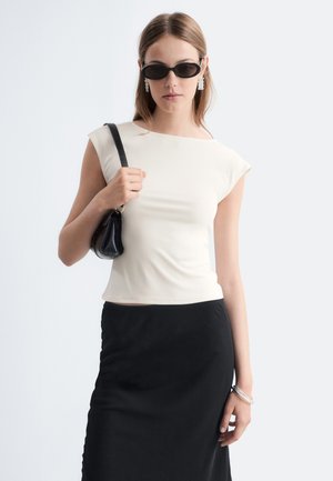 Femme portant des lunettes de soleil noires, un haut blanc sans manches, une jupe noire, tenant un sac à épaule noir et portant des boucles d'oreilles et un bracelet argentés.