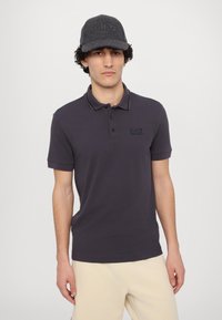 Tmavošedé polo tričko s krátkymi rukávmi a nenápadným logom na hrudi, skombinované s ladiacou tmavošedou baretou.