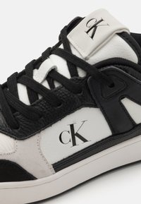 Fekete-fehér tornacipő, bőr és velúr anyagokkal, kiemelkedő "ck" logóval, texturált részletekkel és hagyományos fűzéssel.