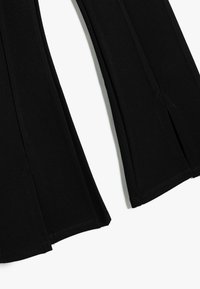Schwarze, ausgestellte Hosen aus weichem Stoff, mit seitlichen Schlitzen in der Nähe des Saums und schlichter Naht. Glatte Textur ohne Muster oder Akzente.