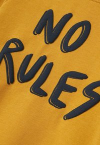 Tela amarilla con un texto grueso y azul oscuro que dice "SIN REGLAS". La textura parece suave con un acabado de letras ligeramente elevadas.