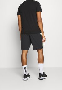 Mies, joka pukeutuu mustaan t-paitaan, mustiin shortseihin neon-yksityiskohdilla, valkoisiin Puma-sukkiin ja mustiin lenkkareihin, seisoo yksinkertaista valkoista taustaa vasten.