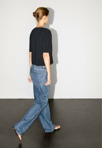 Schwarzes, kurz geschnittenes T-Shirt mit kurzen Ärmeln, kombiniert mit lockeren hellblauen Jeans. Das Modell geht auf einem dunklen Boden vor einem schlichten Hintergrund.