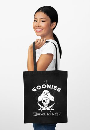 Giovane donna sorridente, che tiene una borsa nera con teschio da pirata bianco, spade incrociate e il testo "The Goonies Never Say Die" su sfondo semplice.