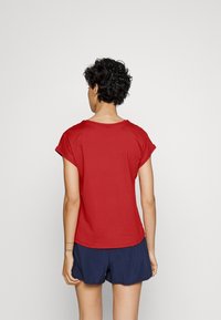 Punainen puuvillainen t-paita, jossa on lyhyet käännetyt hihat, pyöreä pääntie ja rento mitoitus. Vaatetettu tummansinisillä shortseilla, nähty takaa.