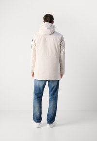 Lys grå vandtæt jakke med hætte, der har en lige pasform, to bagventilationer og en sort detalje på venstre ærme, parret med blå jeans.