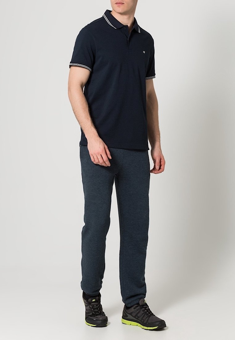 Homme portant un polo bleu marine, un pantalon de survêtement gris foncé et des baskets noires avec des semelles vert fluo, se tenant devant un fond uni.