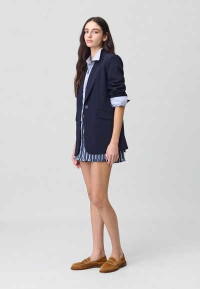 Blazer marine, chemise à carreaux bleus et blancs, jupe rayée avec ourlet à volants. Mocassins en suède marron avec couture décorative.