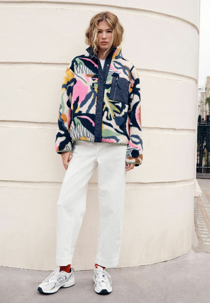 Giacca in pile con design floreale multicolore e tasca blu navy, abbinata a pantaloni bianchi a gamba larga e sneakers grigie.