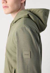 Sudadera con capucha en verde oliva, hecha de tela texturizada. Cuenta con un parche de logo cuadrado en la manga izquierda y un bolsillo frontal.
