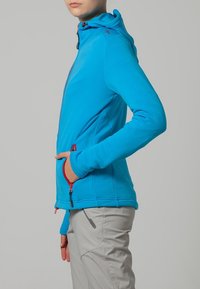Blauwe fleece jacket met een aansluitend ontwerp, rits aan de voorkant en capuchon. Heeft rode details en zijzakken, gecombineerd met lichtgrijze broek.