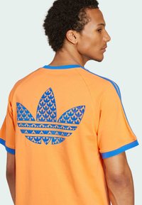 Camisa de manga corta naranja con acentos azules. Presenta un logo gráfico azul en la parte de atrás, que muestra un patrón de hojas y rayas.