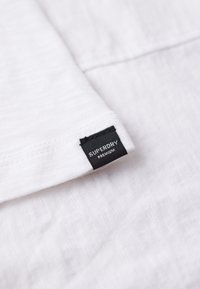 Tela de algodón blanco con un acabado texturizado, que presenta una etiqueta negra "Superdry Premium" cosida en el dobladillo. Diseño simple y limpio.