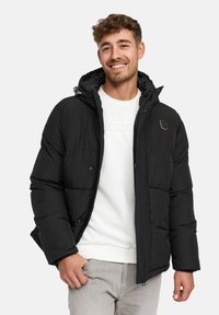 Smilende mand iført sort foret jakke over en hvid sweatshirt og lys grå bukser, stående foran en ensfarvet lys baggrund.