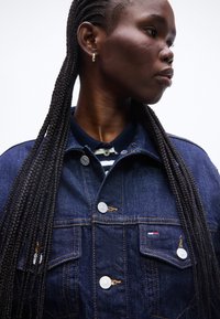 Γυναίκα με μακριά πλεξούδα, που φοράει σκούρο τζιν μπουφάν πάνω από ριγέ πουκάμισο με γιακά, κοιτάζει προς τα πλάγια σε απλό φόντο.