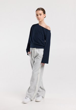 Jonge persoon staand met één hand in de zak, gedragen een marineblauwe off-shoulder sweatshirt, lichtgrijze wijde sweatpants en witte sneakers.