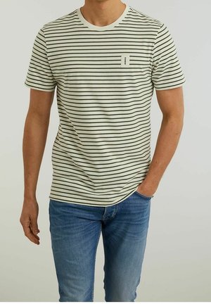 Mann trägt weißes und dunkelgrünes, gestreiftes T-Shirt mit kurzen Ärmeln und Jeans, eine Hand in der Tasche vor einfachem Hintergrund.