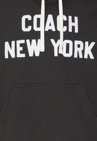 Felpa nera con cappuccio, caratterizzata da grandi scritte bianche "COACH NEW YORK", con lacci e una tasca frontale. Il materiale sembra morbido con una superficie liscia.