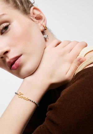 Mujer joven llevando una pulsera dorada "Tous" y dos pendientes, uno con un colgante de dado plateado, posando con la mano cerca del cuello sobre un fondo blanco.