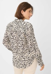 Lange mouwen button-up shirt in crème met zwart gespikkeld patroon. Heeft een puntige kraag, yoke-detail en afgeronde zoom.
