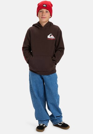 Bruine hoodie met groot logo, rode accenten, voorkant zak. Gepaard met losse blauwe denim broek en zwarte sneakers. Rode gebreide beanie op het hoofd.