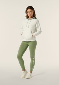 Felpa bianca con cappuccio a righe verdi, abbinata a leggings verde oliva con testo "FREDDY MOV". Sneakers bianche completano l'outfit.