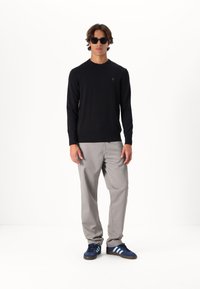 Maglione nero a maglia, pantaloni grigio chiaro e sneakers blu scuro con bande azzurre. Modella che indossa occhiali da sole neri a forma rotonda, in piedi.