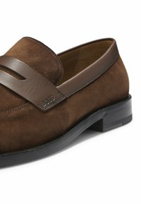 Mocassino uomo in camoscio marrone con dettaglio di cinturino in pelle e suola nera, con marchio in rilievo sottile sul bordo del cinturino.