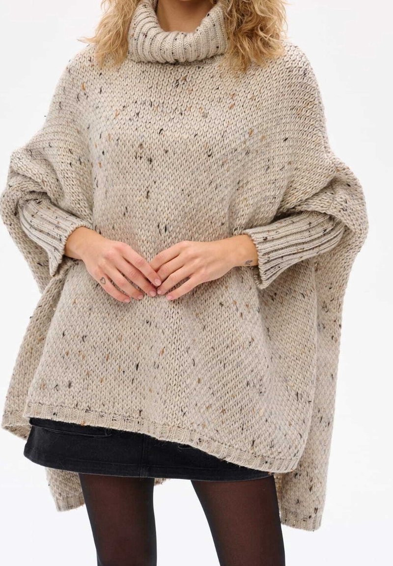 Beige stickad poncho med en bred hög krage, neddragna ärmar och prickig textur. Har en lös passform och ribbade mudd.