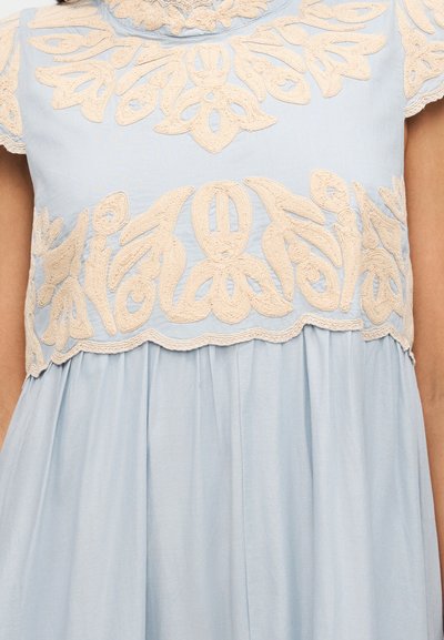 Φωτεινό γαλάζιο φόρεμα με περίπλοκη μπεζ κεντητική λουλουδιών στο στήθος και τα μανίκια, με το ύφασμα να συγκεντρώνεται κάτω από την κεντητική λεπτομέρεια.