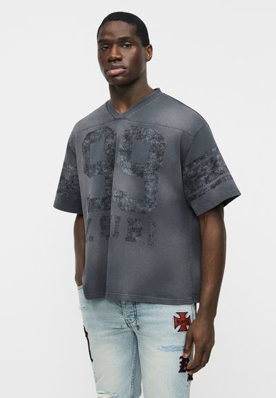 Νεαρός άντρας που φοράει σκούρο γκρι υπερμεγέθη κοντομάνικη μπλούζα με το εκτύπωμα "99" και "ZUBI", συνδυασμένη με ανοιχτό μπλε σκισμένο τζιν με κόκκινα και μαύρα μπαλώματα.