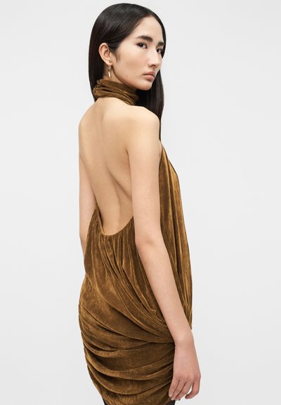 Γυναίκα με μακριά σκούρα μαλλιά, που φοράει καφέ βελούδινο φόρεμα με ανοιχτή πλάτη και πλισέ λεπτομέρειες, στραμμένη στο πλάι μπροστά σε απλό ανοιχτό γκρι φόντο.