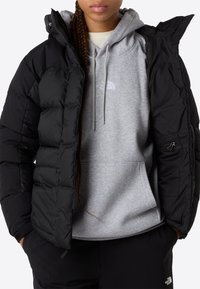 Persona con una chaqueta de plumas negra sobre una sudadera gris de The North Face y pantalones negros, con las manos en los bolsillos.