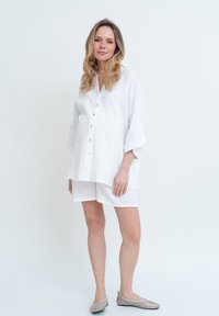 Maglietta bianca oversize, con texture, bottoni frontali e maniche larghe, abbinata a pantaloni corti coordinati. Indossata con scarpe slip-on in rete grigie.
