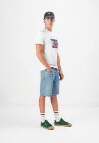 Hvid t-shirt med et navy og rødt grafisk design, lyseblå denimshorts, grønne sneakers med beige såler, grå kasket og stribede strømper.