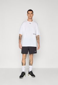 T-shirt blanche surdimensionnée avec un logo Puma noir et du texte. Associé à un short gris foncé et des baskets noires, avec des chaussettes blanches.
