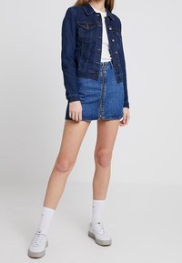 Mörk denimjacka med knappstängning, i kombination med en ljusblå denimkjol med framsidans dragkedja och fransad nederkant, kompletterad med vita sneakers.