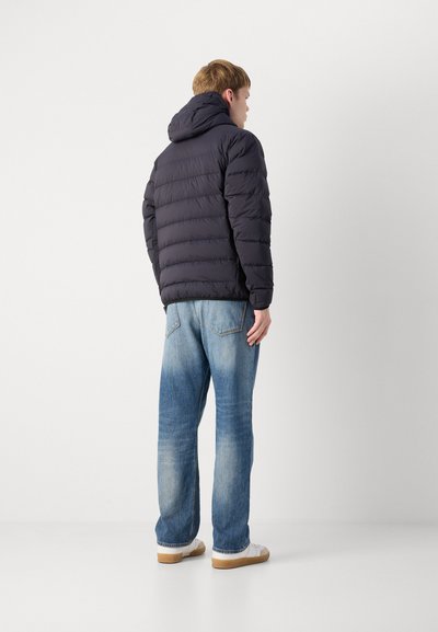 Veste matelassée marine avec une finition texturée, capuche et détails de couture horizontale ; associée à un jean bleu clair et des baskets blanches.