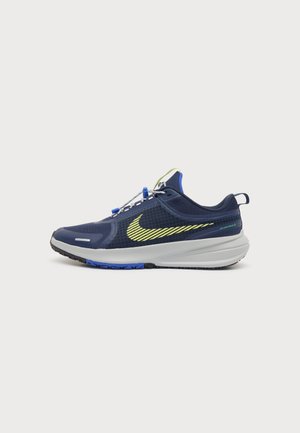 Tumesinine sportlik jalanõu helerohelise Nike logoga, valge tallaga, sinise ja musta haardega ning kiire paelaga süsteemiga ühevärvilisel taustal.
