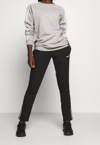 Persona vestida con sudadera gris de Adidas y pantalones de chándal negros de Adidas con rayas blancas, de pie con las manos en los bolsillos contra un fondo liso.