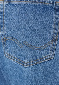 Denim sinised teksad tagantvaates, millel on üks tasku kerge oranži õmblusega ja tasku ümber kaarjas dekoratiivne õmblus.