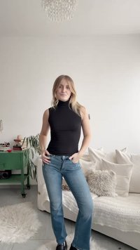Top preto sem mangas com gola alta, jeans azul claro com corte flare e sapatos pretos. O fundo inclui um sofá branco e um armário verde.