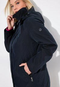 Marinblått vattentätt jacka med huva, frontdragkedja, sidofickor och en logotyp på ärmen. Mjuk textur med minimala designdetaljer.