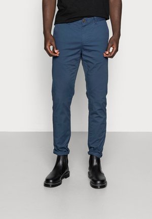 Homme portant un pantalon bleu marine retroussé, des bottines noires et une chemise noire sur un fond clair uni.